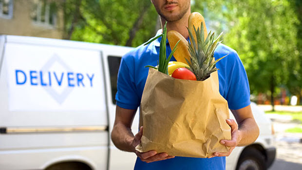 Image of a a man in a blue shirt holding a brown paper bag of groceries. A delivery van is seen in the background.